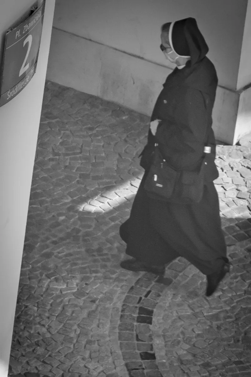 Masked nun in a dark habit walking across a curved cobblestone path